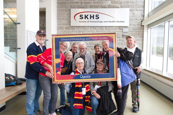 A group of alumni poses behind a picture frame, with the SKHS sign in the background.
